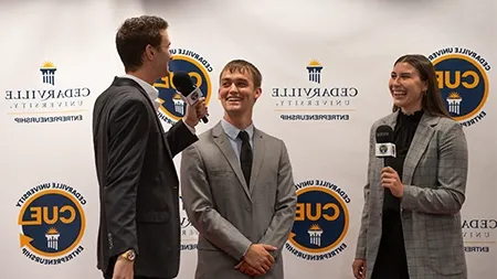 Two students interviewing another student by a backdrop that says 